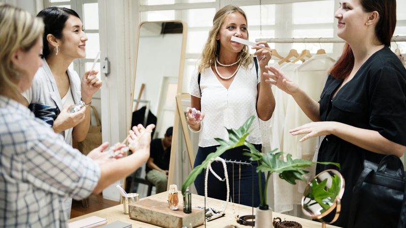 Quatro pessoas, idade média de 35 anos, raça branca, cheirando perfumes em uma loja de moda com roupas e joias sobre a mesa